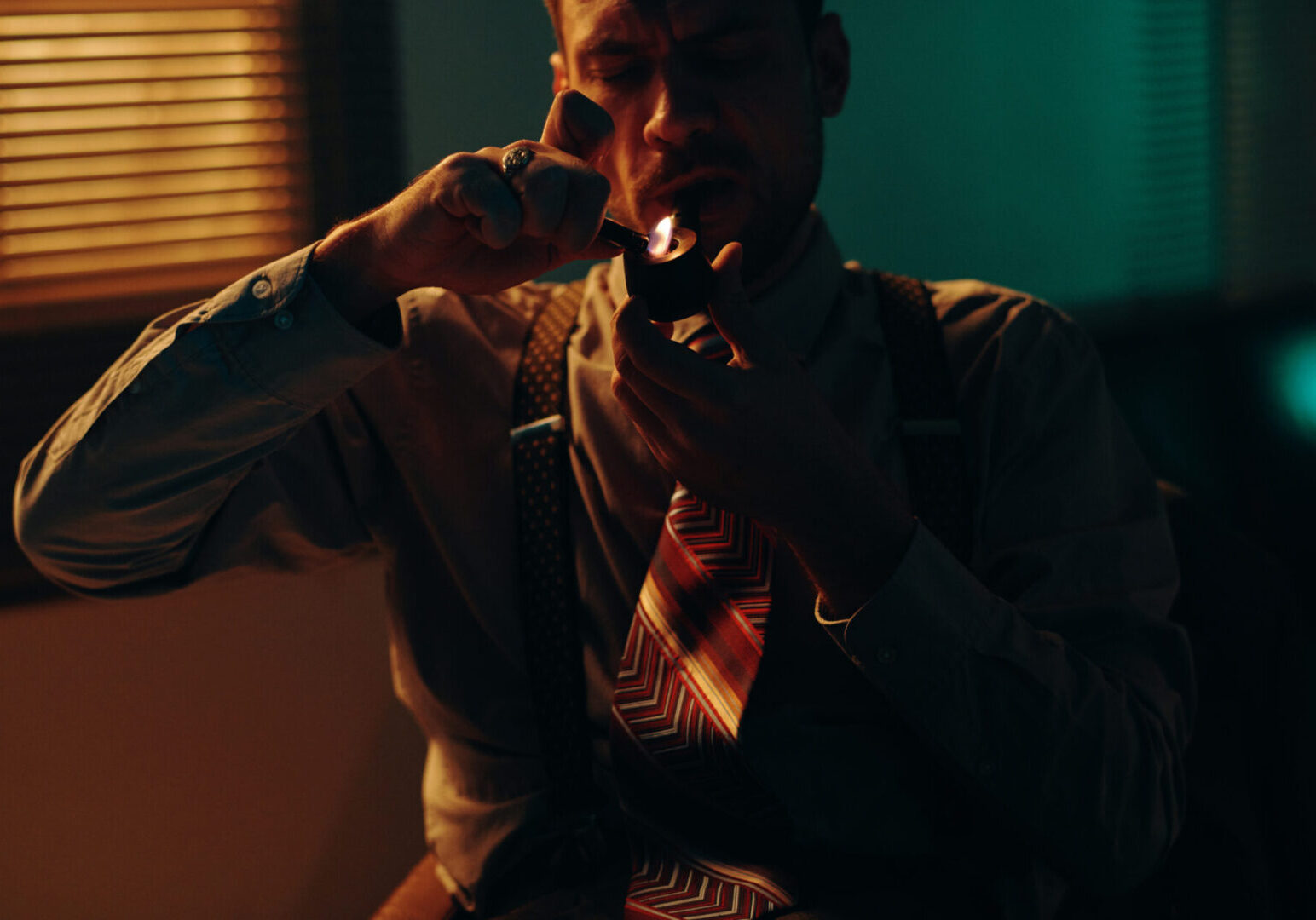Young businessman or police officer in formalwear lighting tobacco pipe while sitting in dark office in front of camera and smoking